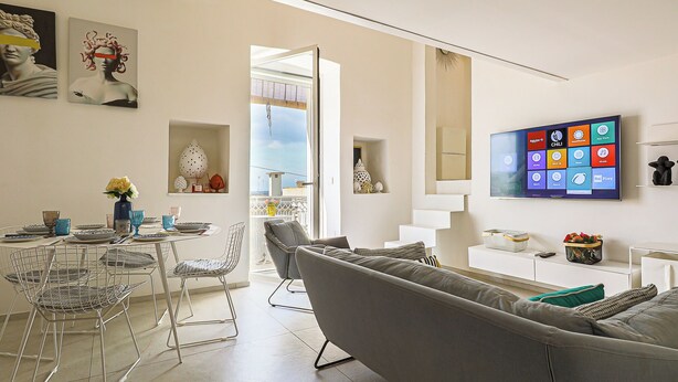 Un soggiorno moderno con un divano grigio, un'area pranzo con un tavolo rotondo e sedie, una TV a schermo piatto montata a parete e un balcone con ringhiera.