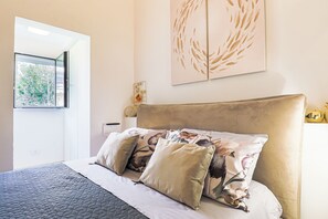 Quarto com cama, almofadas, cabeceira, janela e uma obra de arte na parede.