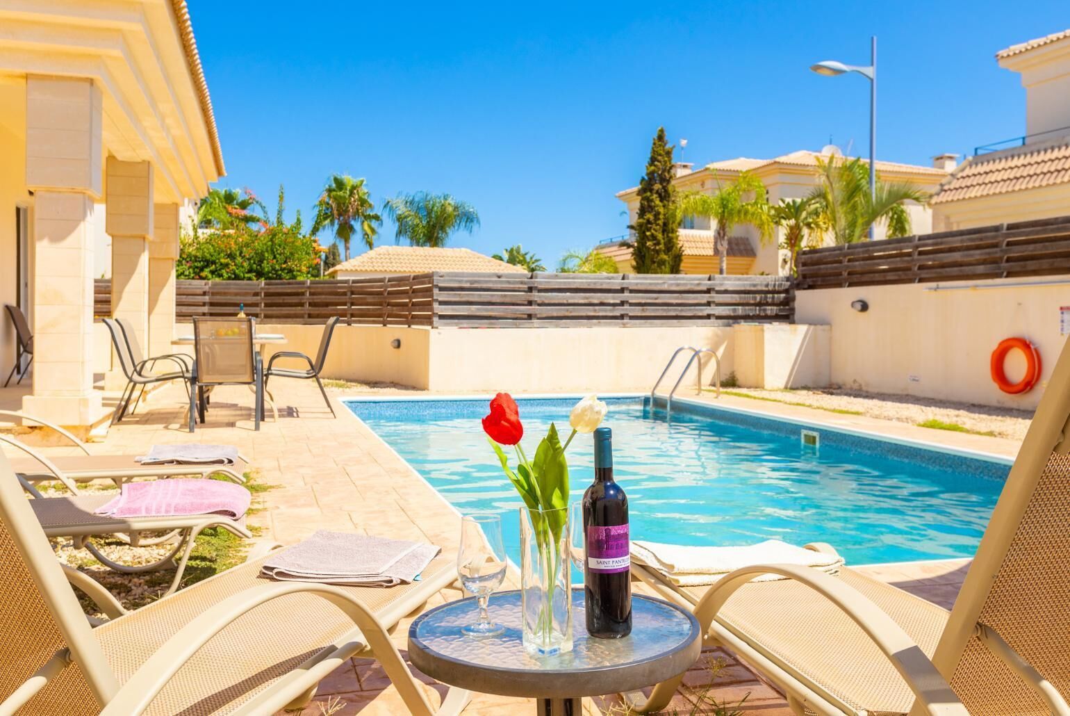 A poolside area with lounge chairs, a table with a bottle of wine and a glass, and a flower vase.