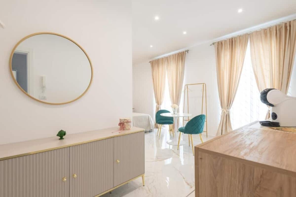 Habitación de hotel moderna con un espejo redondo, un escritorio de madera, un tocador blanco, sillas color aguamarina y una silla con marco dorado.