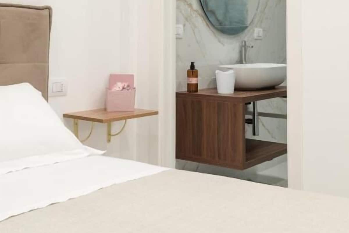 Un dormitorio con una cama, una mesita de noche con un objeto rosa, una mesita auxiliar de madera, y un baño con una pared de mármol, un espejo redondo y un lavabo.