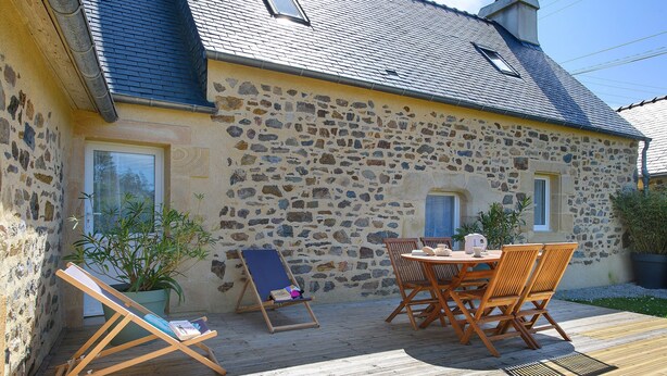 Un bâtiment en pierre doté d’une terrasse en bois, d’une table avec des chaises et d’une chaise longue.