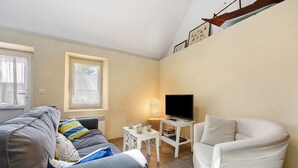 Un salon avec un canapé gris, un fauteuil blanc, une petite table blanche sur laquelle se trouve un téléviseur, et une fenêtre avec des stores.