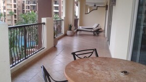En balkong med ett bord och stolar, en krukväxt och utsikt över en swimmingpool och byggnader.