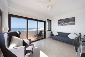 Ein modernes Wohnzimmer mit einer Couch, Sesseln, einem Deckenventilator und einem großen Fenster mit Meerblick.