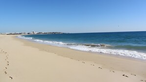 Uma extensa praia de areia com pegadas que levam à água, céu azul límpido e uma linha de costa distante com edifícios.