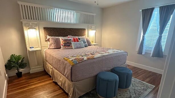 Un dormitorio con cama, lámparas de noche, una mesita de noche, una planta en maceta y una ventana con cortinas.