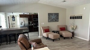 A modern living room with a brown sofa, a dining area with a wooden table and bar stools, a kitchen with dark cabinets, and a 'Beach' artwork on the wall. - 3-bedroom house in tranquil Lynn Haven, beautiful sunsets, & minutes to PCB. (Lynn Haven)