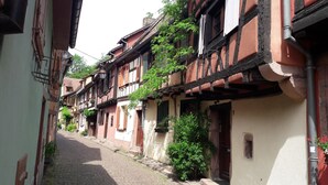 Une ruelle étroite pavée, bordée de bâtiments traditionnels à colombages, chacun doté d’un petit balcon et d’une porte visible.