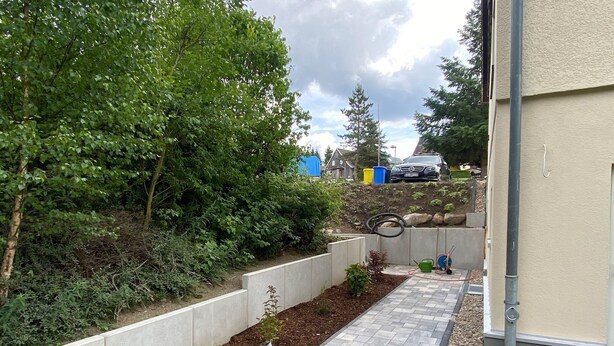 Ein gepflasterter Weg führt zu einem Gebäude, gesäumt von einem erhöhten Blumenbeet mit Pflanzen, einer Betonretmlaug und einer Metallrohrleitung.