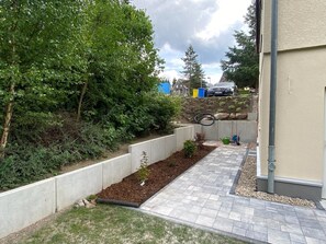 A paved pathway leading to a building, bordered by a raised garden bed with plants, a concrete retaining wall, and a metal pipe. - Oberharzruhe Vacation Home - "Fuchsbau" Basement Unit for 2 People, Covered Terrace (Oberharz am Brocken)