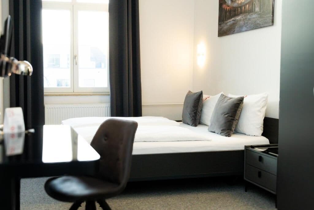 A hotel room with a bed, a desk, a chair, a nightstand, and a window with curtains.