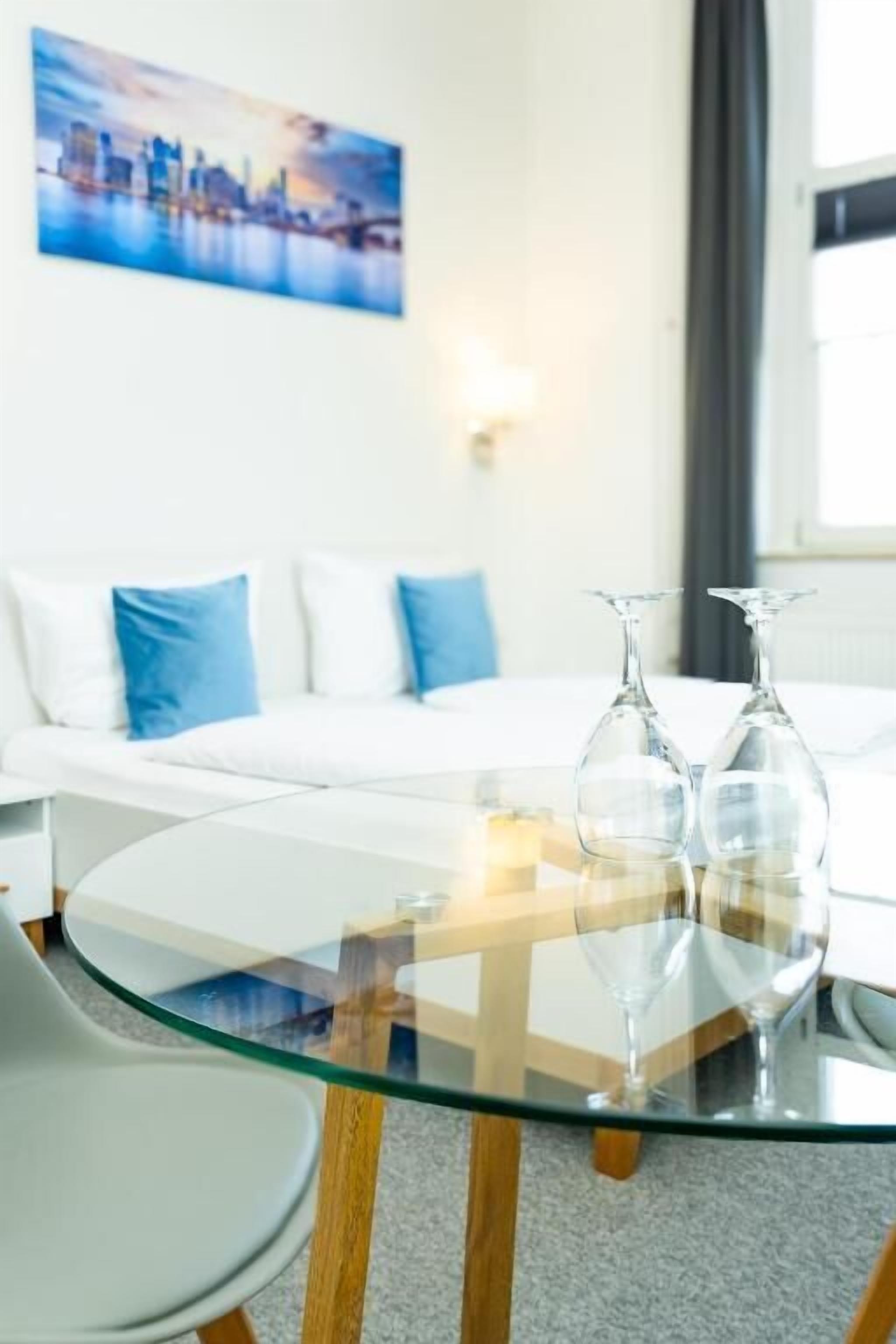 A modern hotel room with a glass-top dining table, a white sofa with blue cushions, and a cityscape painting on the wall.