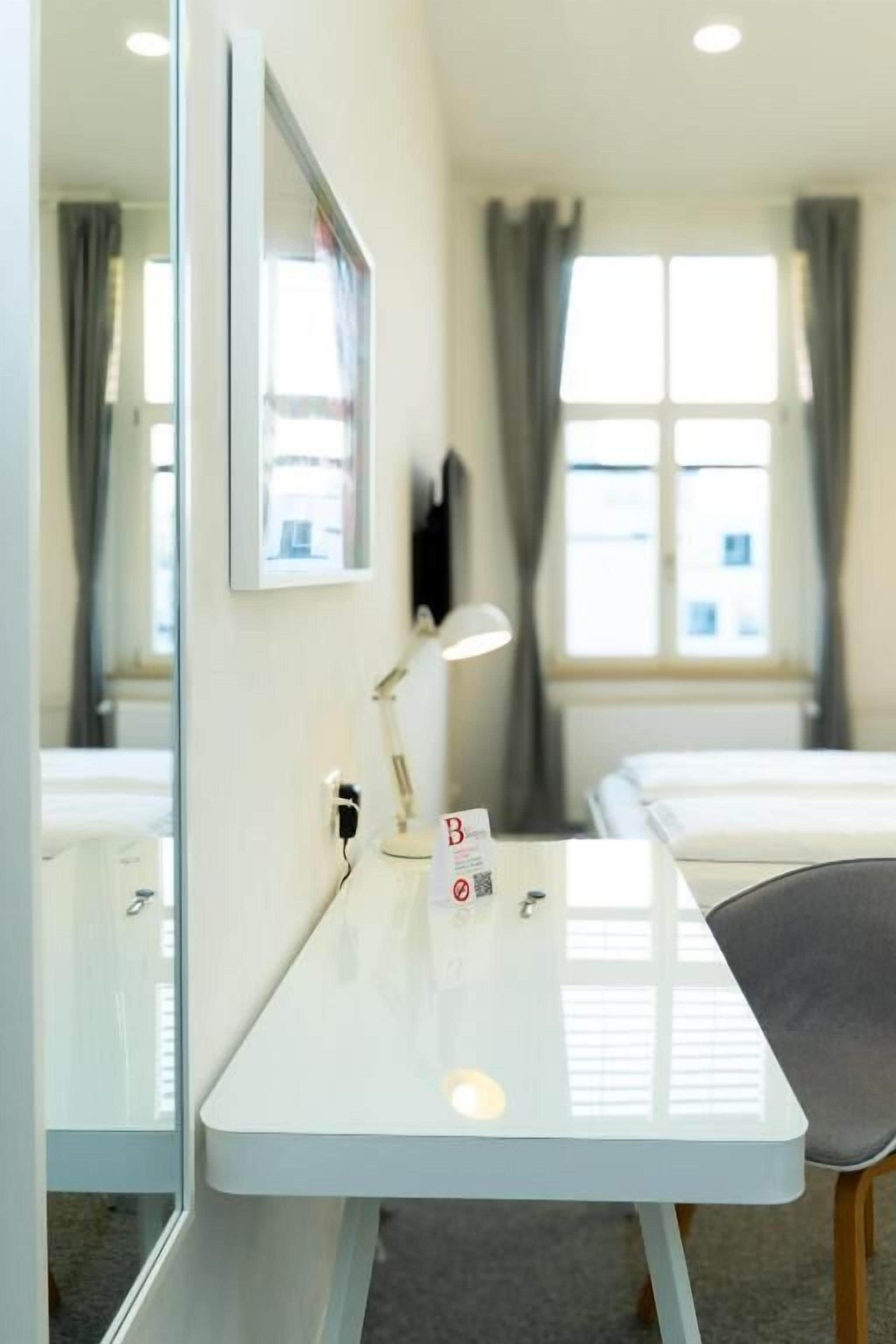 Une chambre d’hôtel avec un lit, un bureau avec une lampe, un miroir et une fenêtre avec des rideaux.