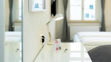 Une chambre d’hôtel avec un lit, un bureau avec une lampe, un miroir et une fenêtre avec des rideaux.