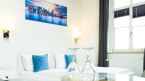 Ein Hotelzimmer mit einem weißen Sofa, einem Glastisch mit Weingläsern, einem Stadtbild als Gemälde und einem Fenster mit Jalousien.