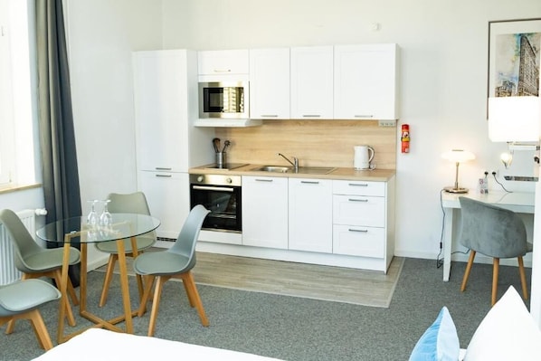 Cozinha compacta com armários brancos, micro-ondas e pia. Há uma área de jantar com mesa de vidro e cadeiras, e uma pequena escrivaninha com luminária.