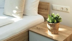 Ein ordentlich bezogenes Bett mit weißer Bettwäsche, ein Nachttisch mit einem gewebten Korb und eine Wandleuchte.