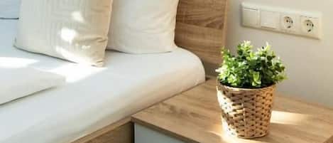 Una cama bien hecha con ropa de cama blanca, una mesita de noche con una cesta tejida y una lámpara de pared.