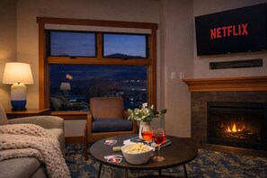 Una habitación de hotel acogedora con chimenea, un televisor montado en la pared, un sillón cómodo, una mesa con aperitivos y bebidas, y vistas a la ciudad al atardecer.
