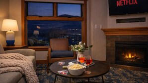 Camera d'albergo accogliente con camino, televisore a parete, una poltrona comoda, un tavolo con snack e bevande e vista sulla città al crepuscolo.
