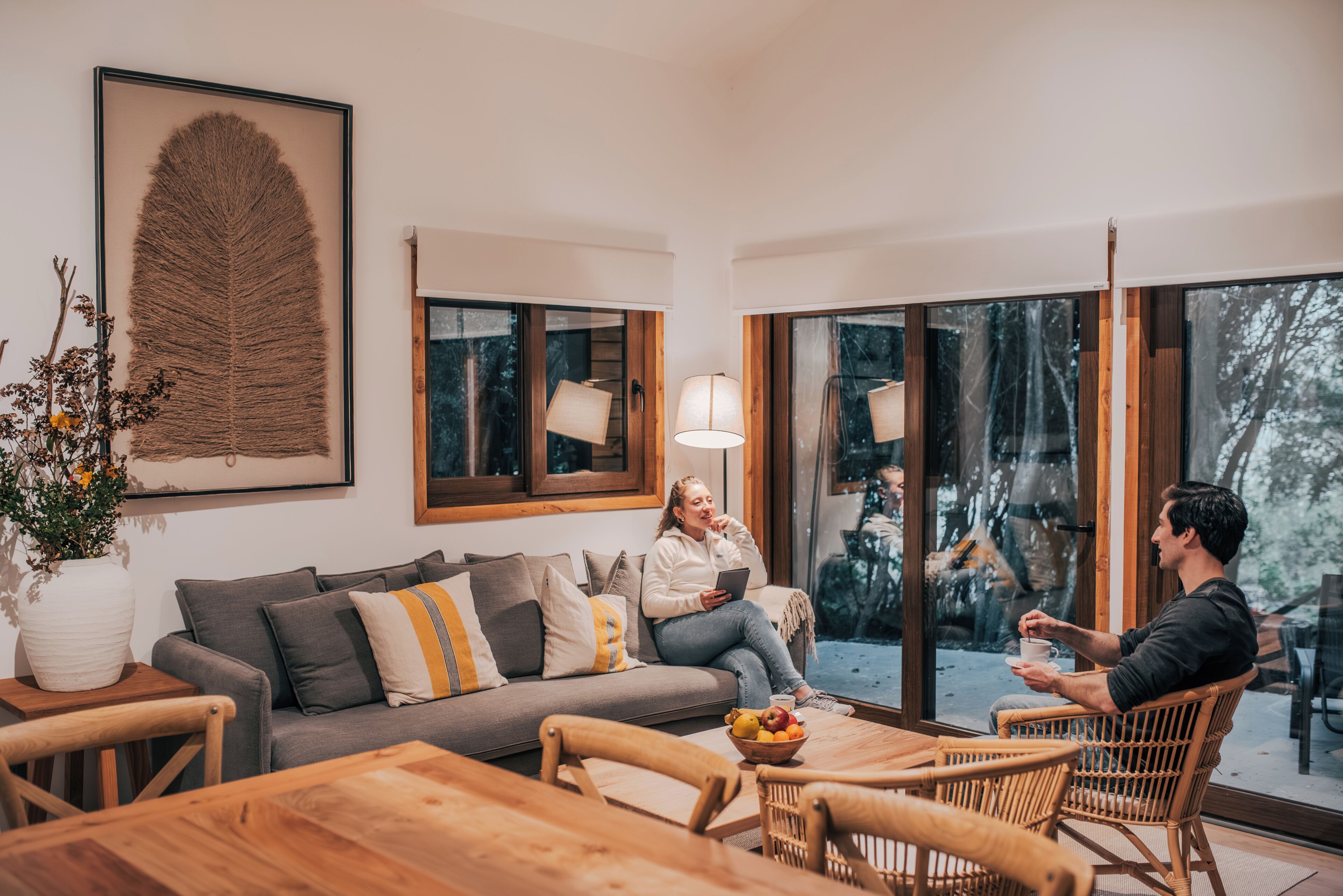 A modern living room with a sofa, a dining table, and chairs. There is a large window with a view of trees.