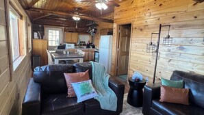 Interior acolhedor de uma cabana com teto de madeira, sala de estar com sofá e mesa de centro, espaço de cozinha com geladeira e micro-ondas, e mesa de jantar.