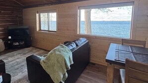 Interior de uma cabana com teto de madeira, um sofá, uma mesa e vista para a água.