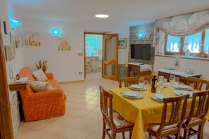 Ein Essbereich mit gelbgrünem Tischlein, Holzstühlen und einem Fernseher an einer Steinwand.