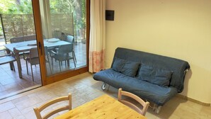 Une pièce avec une table en bois, un canapé bleu et une porte-fenêtre coulissante donnant sur une terrasse extérieure équipée d’une table et de chaises.