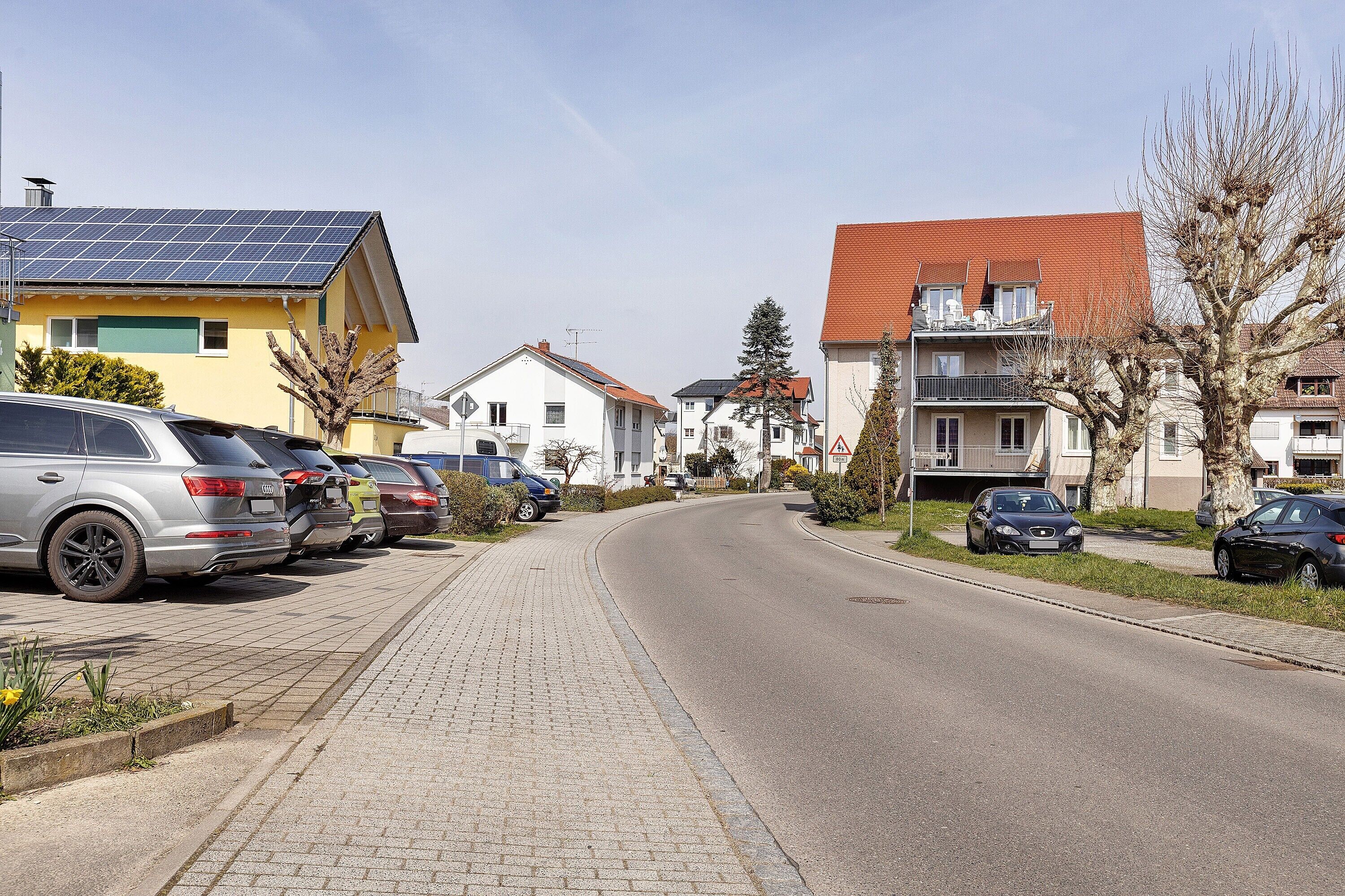 A residential street with parked cars, houses with solar panels, and a clear sky.