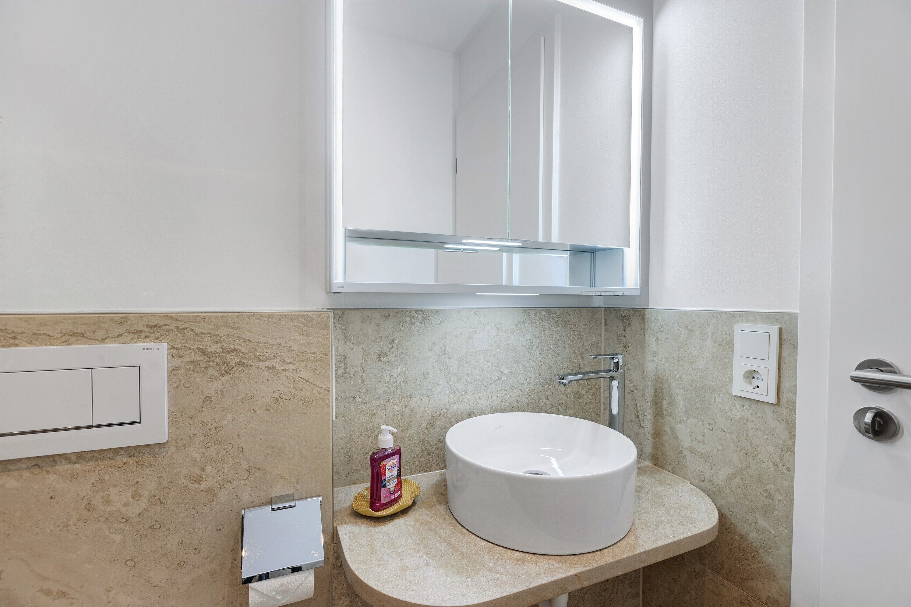 A bathroom with a round sink, a mirror, a soap dispenser, and a towel warmer.