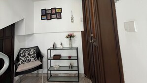 Ein Zimmer mit Holztür, einem schwarzen Stuhl mit gemustertem Kissen, einer Glastrennwand mit Dekorationsgegenständen, einer Wandleuchte und einer Bildergalerie in Rahmen.