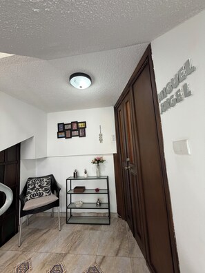A room with a wooden door, a black chair with a patterned cushion, a glass shelf with decorative items, a wall-mounted light fixture, and a framed picture collection. - Charming & Cozy Suite in Quito with Great Location (Quito)