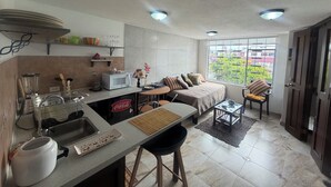 Eine kompakte Küche mit Mikrowelle, Spüle und Mixer, ein Essbereich mit Sofa und Stühlen sowie ein großes Fenster mit Blick auf Grünflächen.