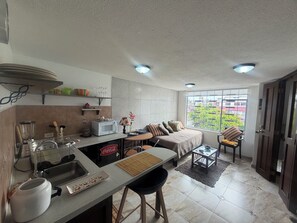 A compact kitchen with a microwave, sink, and blender, a dining area with a sofa and chairs, and a large window offering a view of greenery. - Charming & Cozy Suite in Quito with Great Location (Quito)