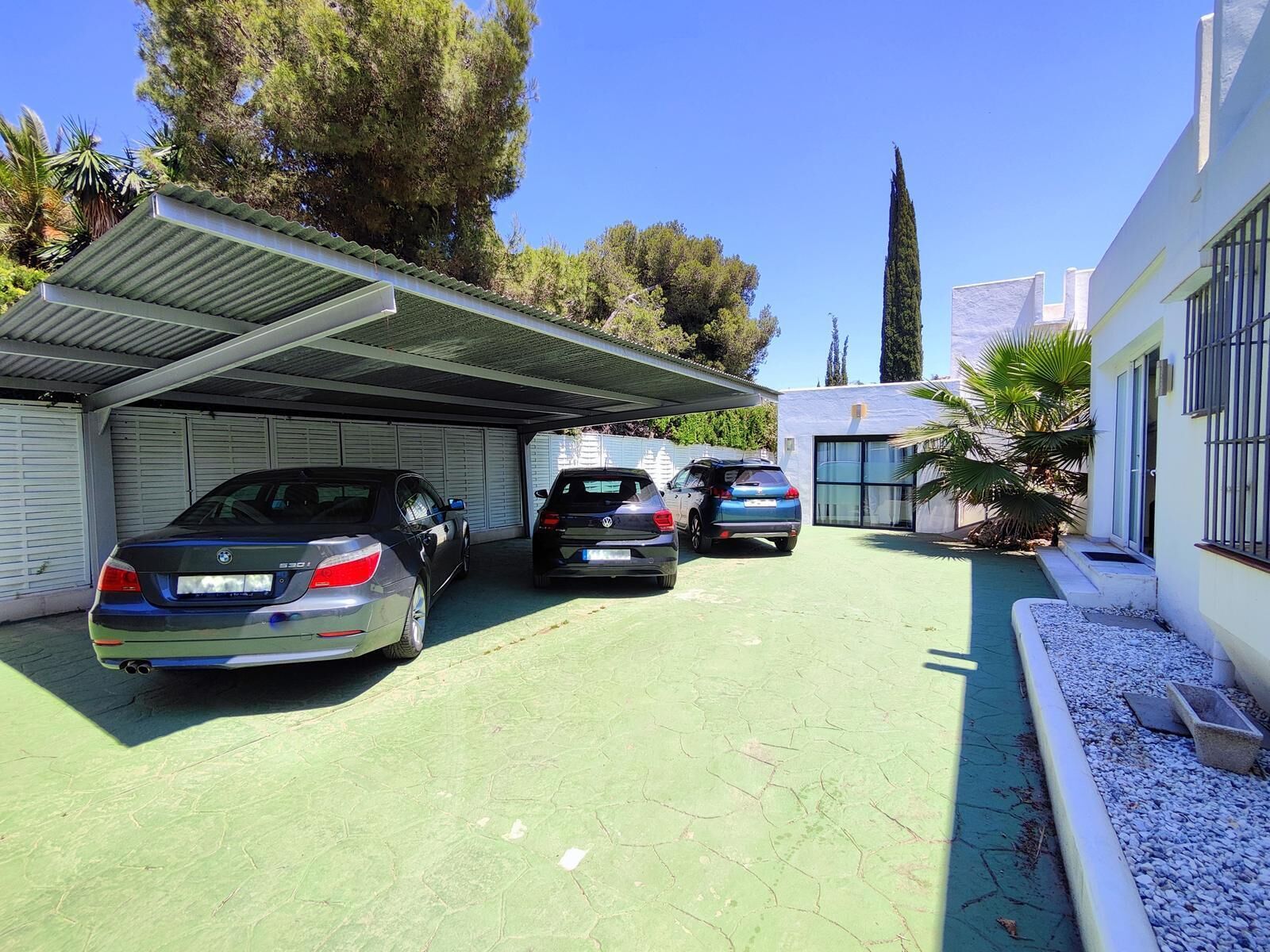 A covered parking area with three cars parked, a building with a white facade, and a tall tree in the background.