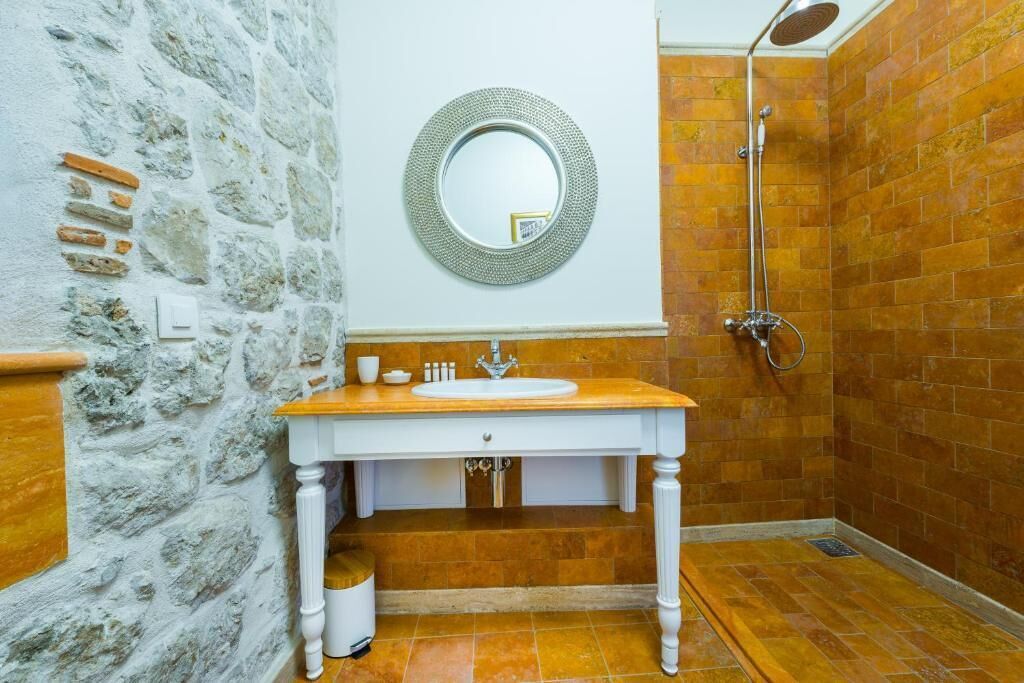 A bathroom with a stone wall, a wooden vanity, a round mirror, and a shower area.