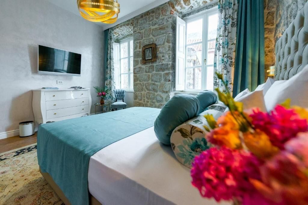 A bedroom with a stone wall, a bed with a teal bedspread, a television on the wall, and a window with curtains.