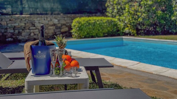 A poolside setting with a bottle of champagne, a pineapple, and oranges on a table. - Villa Camilla: Private Pool & BBQ, Panoramic Garden Near Le Saline beach (Punta de Su Turrione)