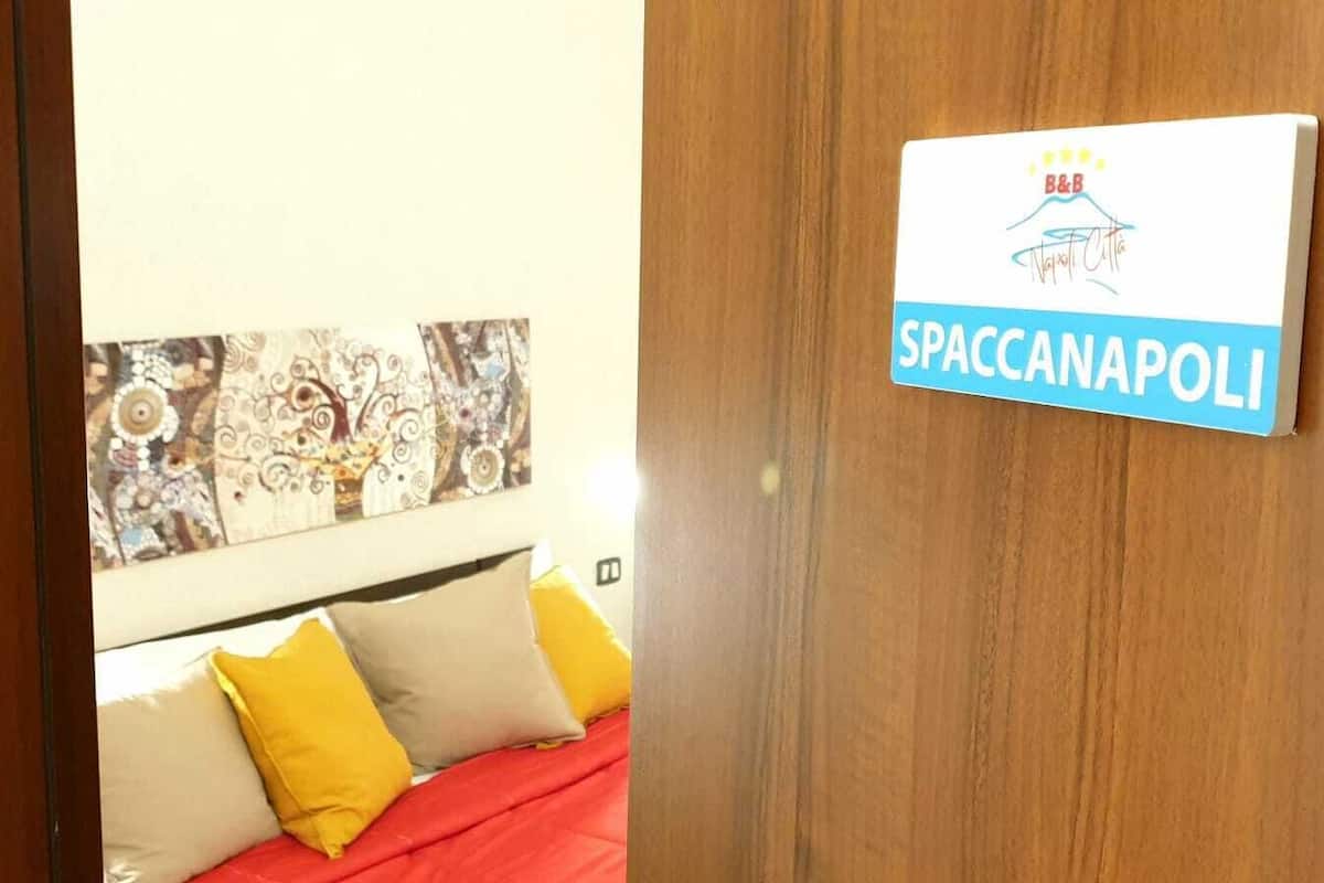 Hotellihuone, jossa on sänky, koristekuvioitu seinä ja kyltti, jossa kerrotaan, että paikka on Spaccanapoli.