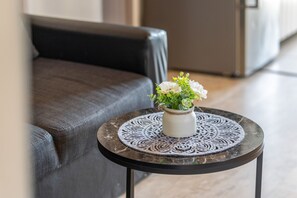 Une table ronde ornée d’un plateau décoratif, un vase blanc rempli de fleurs et, au fond, un canapé gris.