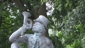 Una estatua de una figura sentada con una trompeta, ubicada sobre un borde de concreto con una barandilla metálica al fondo.