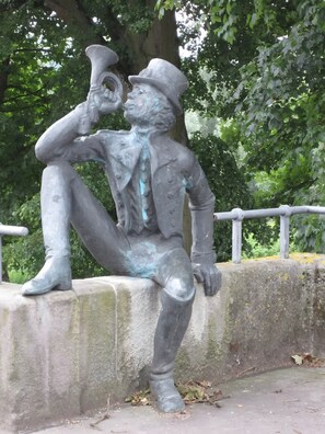 Une statue représentant une figure assise tenant une trompette, placée sur une corniche en béton avec une rambarde métallique en arrière-plan.