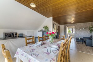 Un coin repas avec une table dressée pour quatre personnes, des chaises en bois, un vase avec des fleurs et un espace cuisine équipé d’un grille-pain Samsung.