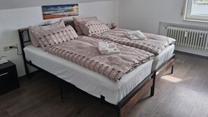 Un lit double avec un cadre en bois, un matelas blanc et des oreillers assortis. Il y a un radiateur au mur et une fenêtre avec des stores.