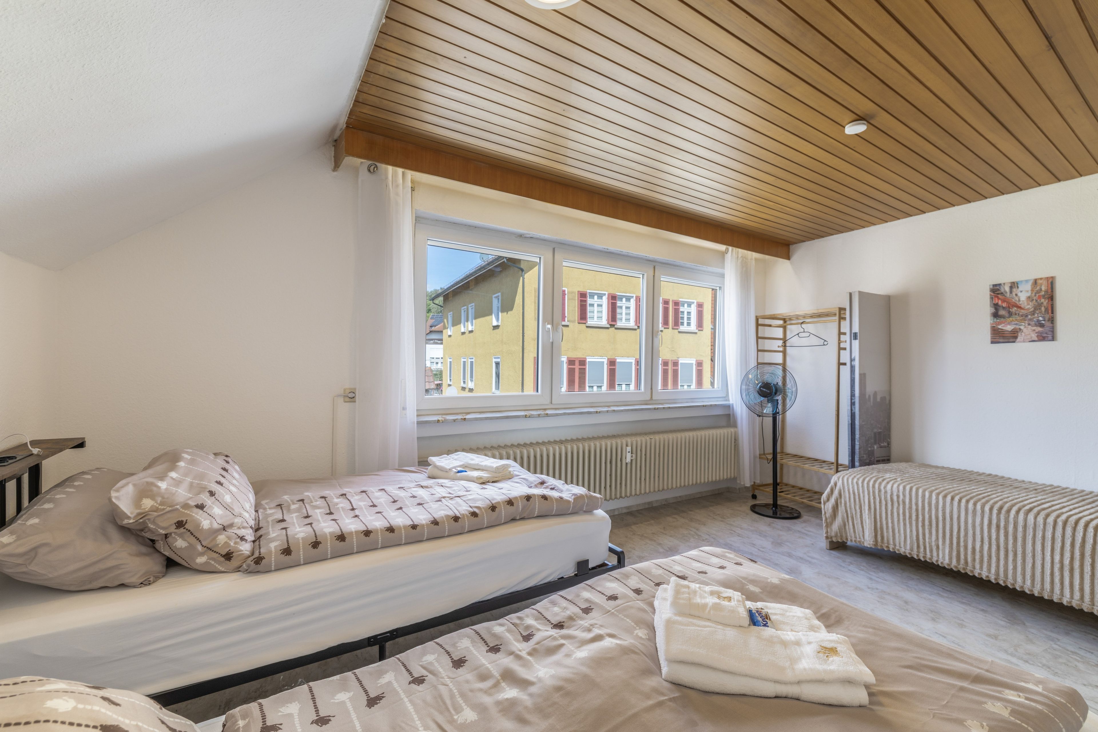 A bedroom with a bed, a bedside table, a radiator, a window, and a wall-mounted fan.