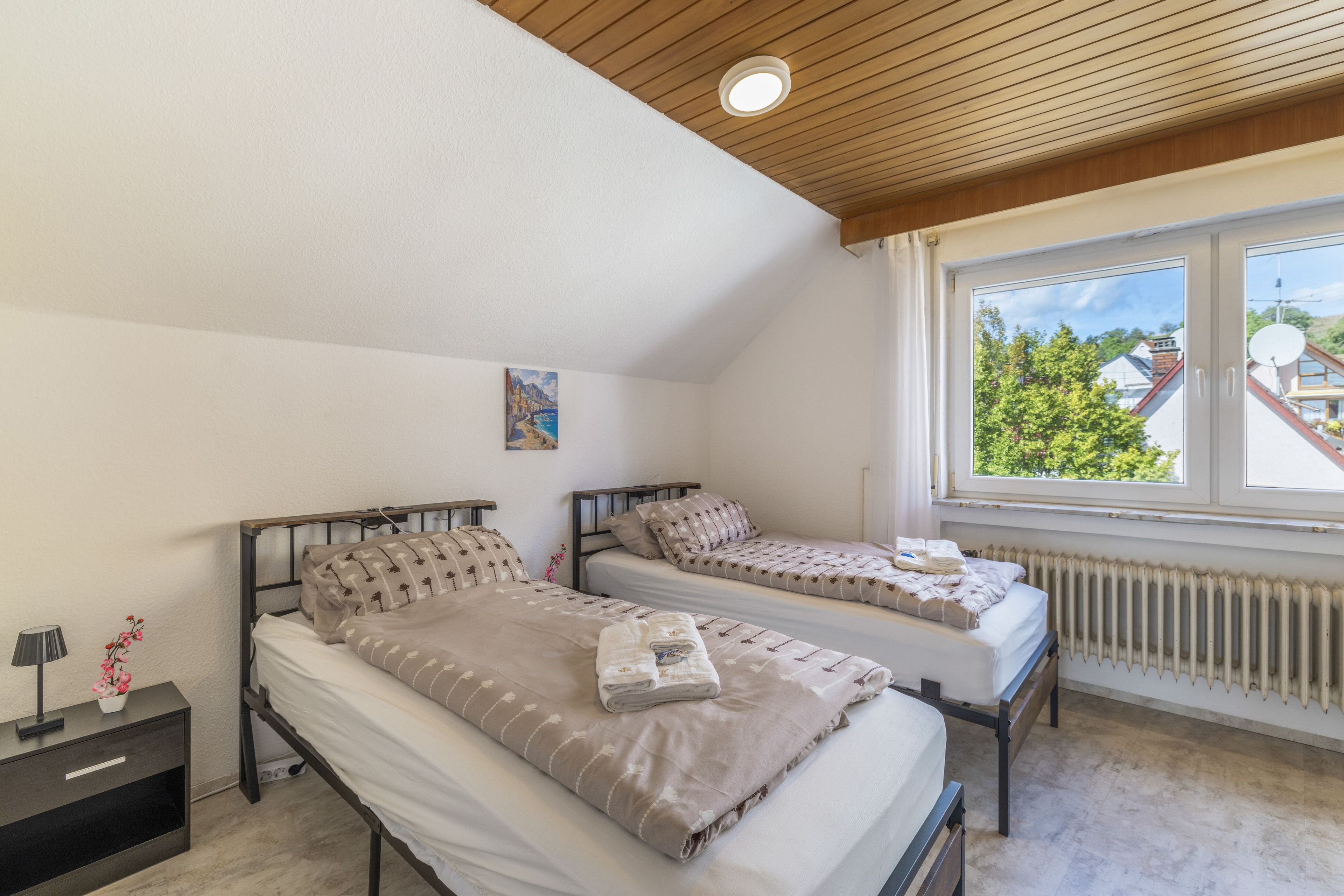 Una camera da letto con due letti singoli, un comodino, una lampada, un quadro e una finestra con vista sulle case.