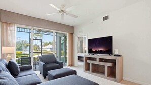 Een moderne woonkamer met een flatscreen-tv, grijze banken, een plafondventilator en uitzicht op het buiten.