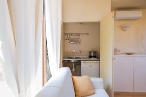 Una cocina compacta con un lavamanos, estufa y lavavajillas, un sofá blanco con un cojín color beige, y una ventana con cortinas.